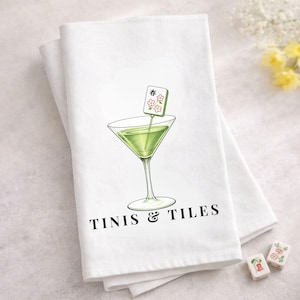 Puede incluir: Pa&ntilde;o de cocina blanco con una ilustraci&oacute;n de c&oacute;ctel verde. La copa de c&oacute;ctel tiene una guarnici&oacute;n con la palabra "春" y flores. El texto "TINIS & TILES" est&aacute; impreso debajo. Dos fichas de mahjong est&aacute;n en la esquina inferior derecha.