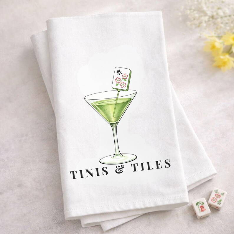Puede incluir: Pa&ntilde;o de cocina blanco con una ilustraci&oacute;n de c&oacute;ctel verde. La copa de c&oacute;ctel tiene una guarnici&oacute;n con la palabra "春" y flores. El texto "TINIS & TILES" est&aacute; impreso debajo. Dos fichas de mahjong est&aacute;n en la esquina inferior derecha.
