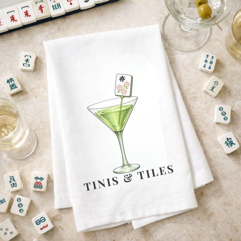 May include: White kitchen towel with a martini glass illustration and the text "TINIS & TILES". The martini glass contains a green liquid and a decorative garnish. Mahjong tiles are scattered around the towel.