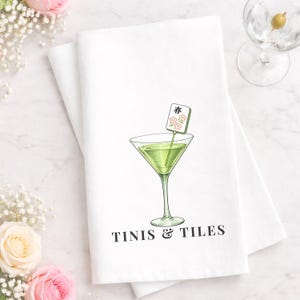 May include: White kitchen towel with a martini glass illustration and the text "TINIS & TILES". The martini glass contains a green liquid and a decorative garnish. Mahjong tiles are scattered around the towel.