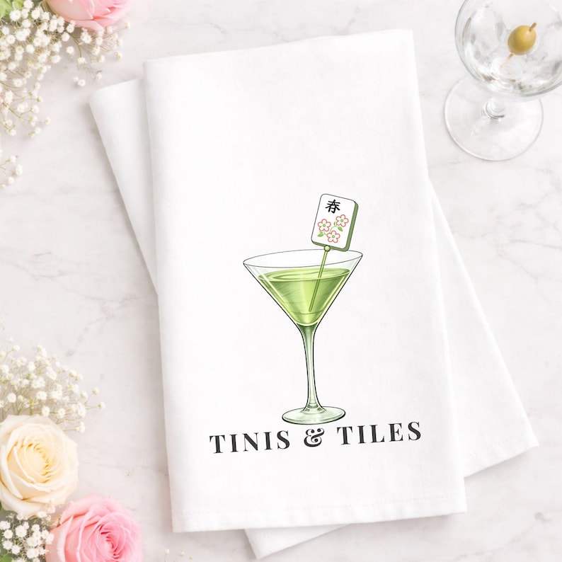 May include: White kitchen towel with a martini glass illustration and the text "TINIS & TILES". The martini glass contains a green liquid and a decorative garnish. Mahjong tiles are scattered around the towel.