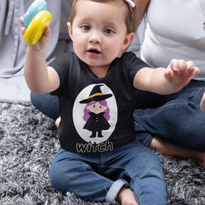 Puede incluir: Un bebé con una camiseta negra con un diseño de bruja y la palabra "witch" impresa debajo. El bebé sostiene un juguete azul y amarillo. El bebé lleva vaqueros azules.
