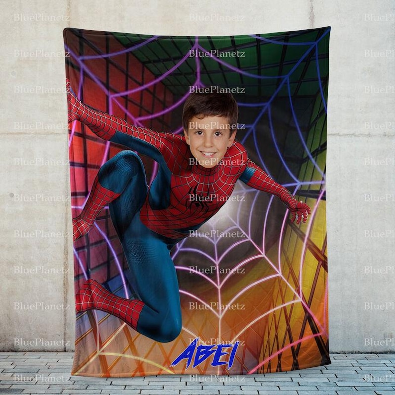 May include: A fleece blanket with a colorful spiderweb design. A young boy dressed as Spiderman is hanging from the web. The blanket has the name "ABEI" printed on it in blue.