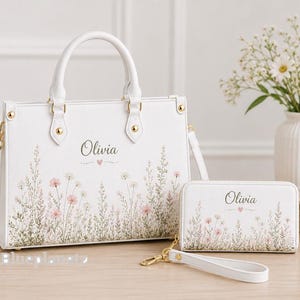 Puede incluir: Un bolso blanco y una cartera rectangular a juego, ambos con un diseño estampado de delicadas flores silvestres rosas y verdes en la base. El nombre Olivia está impreso en una fuente cursiva oscura sobre un pequeño corazón.