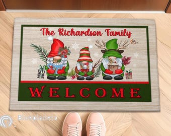 Può includere: Un tappetino di benvenuto con sfondo in legno presenta tre gnomi con cappelli rossi e tazze in mano. Il testo "The Richardson Family" è sopra gli gnomi e la parola "WELCOME" sotto.