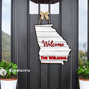 Può includere: Un cartello di benvenuto in legno bianco a forma dello stato della Georgia. Il cartello ha un fiocco rosso in cima e dice "Welcome The Wilsons" in lettere rosse.