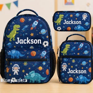 May include: A dark blue backpack, lunch bag, and pencil case set featuring a space-themed pattern with dinosaurs, rockets, planets, and sports balls. Each item is personalized with the name Jackson in white text against the navy background.