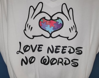 May include: White t-shirt with a black and white image of two hands forming a heart shape with a colorful puzzle piece heart inside. The text "LOVE NEEDS NO WORDS" is printed below the hands.