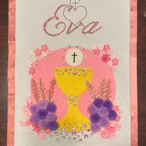 May include: A white fabric banner with a pink border and pink and purple felt flowers. The banner has a yellow chalice with rhinestones, a pink circle with a cross, and the name "Eva" in pink glitter. The date "May 3, 2025" is written in pink glitter at the bottom.