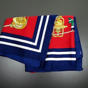 May include: A folded silk scarf with a nautical theme. The scarf features a red background with gold and white nautical illustrations, bordered by a navy blue frame with white stripes. The scarf is folded, showing the design on the front.