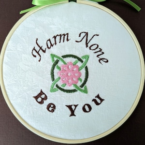 May include: Embroidered hoop art with the words "Harm None Be You" in brown thread. A central design features a pink flower within a green Celtic knot. The fabric is a light cream color, and the hoop is made of wood. A green ribbon is tied at the top.