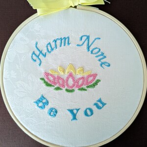 May include: White embroidered hoop with the text "Harm None Be You" in blue thread. A pink and yellow embroidered flower is in the center of the hoop.