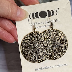 May include: A pair of gold-toned earrings with a detailed, intricate design. The earrings are round and have a hook closure. The earrings are handcrafted in California and are made by Urban Moon.