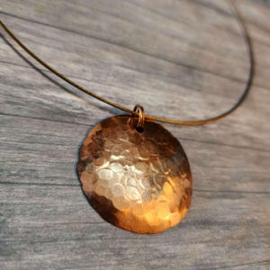 May include: A circular copper pendant with a hammered, textured surface that reflects light. The pendant is suspended from a thin, dark, flexible wire necklace that forms a wide arc, creating a simple and clean jewelry piece.