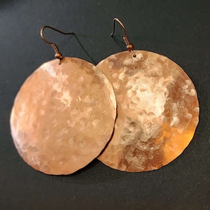 May include: A pair of round, copper-coloured earrings featuring a hammered, textured surface. Each circular disc is suspended from a dark metal hook wire, creating a dangling design that reflects light across the uneven, metallic finish of the metal.
