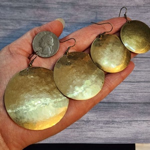 May include: Three pairs of gold-toned hammered metal earrings with a circular shape. The earrings are shown in a hand, with the largest earring being the size of a quarter.