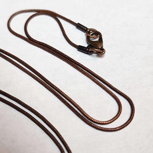 May include: A close-up of a brown necklace with a lobster clasp. The necklace is made of a thin, woven cord. The clasp and the ends of the cord are a darker brown color. The background is a plain white surface.