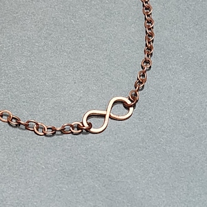 May include: A copper-colored metal chain necklace featuring a central infinity symbol pendant. The chain consists of small, uniform circular links connected to the sides of the infinity loop, creating a continuous, delicate, and metallic jewelry piece.