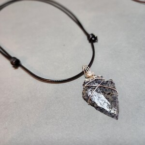 May include: A necklace featuring a dark gray and black speckled stone arrowhead pendant wrapped in thin silver-colored wire. The pendant hangs from a smooth black cord that includes two adjustable sliding knots for length customization.