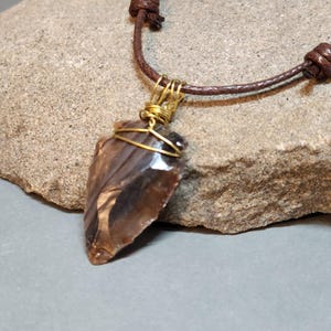 May include: A brown arrowhead pendant with a translucent, layered appearance, secured by gold-colored wire wrapping. The pendant hangs from a dark brown cord necklace featuring adjustable sliding knots for a customizable fit around the neck.