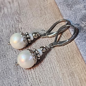 May include: A pair of leverback earrings featuring large, iridescent white pearl beads. The pearls are accented with silver-toned metal floral and bead details. The earrings are displayed on a wooden surface.