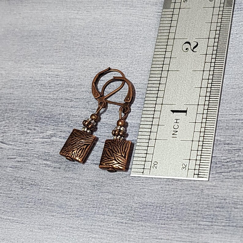 Op de afbeelding: Koperkleurige oorbellen met een rechthoekige hanger met een bladerenpatroon. De oorbellen zijn ongeveer 2,5 cm lang, met een oorhaaksluiting en kleine kraalaccenten. Een liniaal ligt naast de oorbellen.