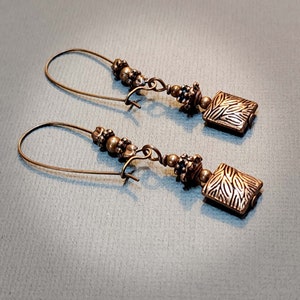 May include: A pair of gold-toned dangle earrings with a square-shaped charm featuring a textured design. The earrings are made with small beads and a wire hook.
