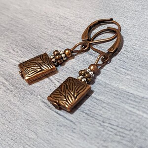 Op de afbeelding: Een paar koperkleurige oorbellen. Elke oorbel heeft een rechthoekige hanger met een bladerpatroon, verbonden met een klapbeugel. Kleine kralen in zilver- en kopertinten accentueren het ontwerp. De oorbellen worden gepresenteerd tegen een grijze achtergrond.