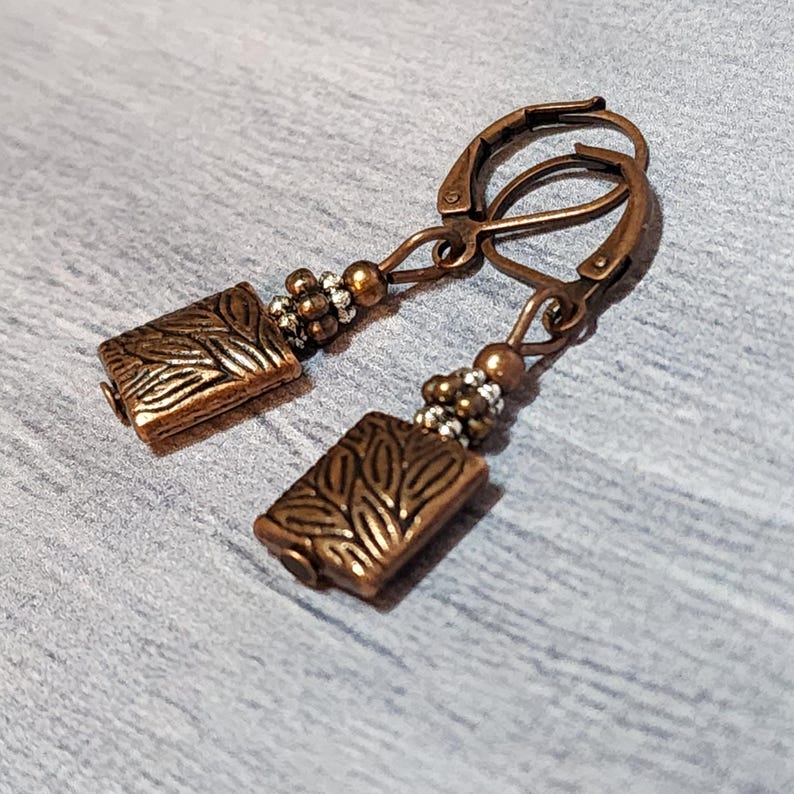 Op de afbeelding: Een paar koperkleurige oorbellen. Elke oorbel heeft een rechthoekige hanger met een bladerpatroon, verbonden met een klapbeugel. Kleine kralen in zilver- en kopertinten accentueren het ontwerp. De oorbellen worden gepresenteerd tegen een grijze achtergrond.