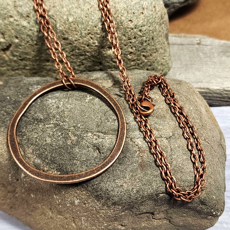 Op de afbeelding: Een koperen ketting met een grote, ronde hanger. De hanger is gemaakt van gehamerd koper en heeft een rustieke, organische uitstraling. De ketting is ook van koper en heeft een delicaat, schakellink-ontwerp.