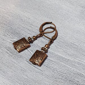 Op de afbeelding: Een paar koperkleurige oorbellen met rechthoekige, met bladeren bewerkte hangers. De oorbellen hebben een klapsluiting en zijn versierd met kleine kralen. De sieraden worden gepresenteerd op een grijze ondergrond.