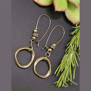 May include: A pair of antique brass earrings with a hoop design. The earrings have a long hook and are adorned with small beads. The earrings are displayed on a dark surface with green foliage in the background.