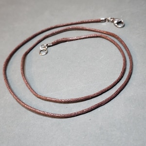 May include: A brown cord necklace featuring a braided texture and a silver-toned metal lobster claw clasp at one end, with a matching silver-toned metal jump ring at the other end to secure the cord for use as a jewelry component.