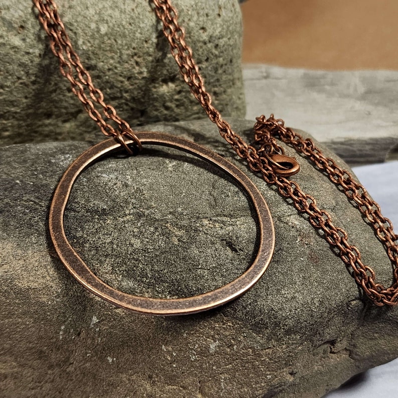 Op de afbeelding: Een koperen ketting met een grote, ronde hanger. De hanger is gemaakt van dik, gehamerd koper en heeft een rustieke, organische uitstraling. De ketting is ook van koper en heeft een eenvoudig, schakellink-ontwerp.