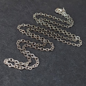May include: A silver-colored chain necklace with an oval link design. The necklace is displayed on a dark surface. The chain is composed of multiple connected links, and a clasp is visible at one end.