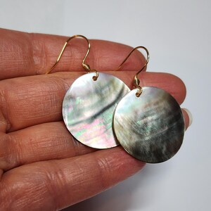 May include: A pair of iridescent, round earrings. Each earring features a circular, mother-of-pearl disc with a shimmering, pearlescent surface. The earrings have gold-colored, curved ear wires. The earrings are held in a hand.