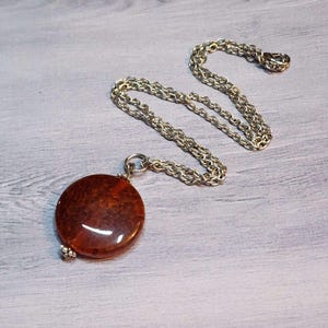 May include: A round, reddish-brown pendant necklace with a silver-colored chain. The pendant has a textured surface and a small silver-colored bead at the bottom. The chain is a classic link style.