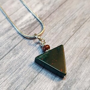 May include: A triangular pendant necklace with a dark green stone, featuring red speckles, hangs from a silver chain. A small, round, brown bead accents the top of the pendant. The necklace rests on a grey wooden surface.