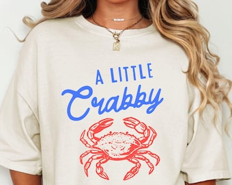 以下が含まれることがあります： 赤色のカニのイラストの上に、青い筆記体で「A Little Crabby」と書かれたアイボリー色のTシャツ。 ゆったりとしたフィット感で、カジュアルウェアに最適です。 シンプルで人目を引くデザインです。