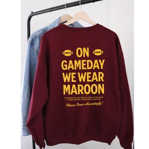 May include: Maroon sweatshirt with the text "ON GAMEDAY WE WEAR MAROON" in yellow. The text is above the phrase "Please Dress Accordingly!". A denim jacket hangs on the rack behind the sweatshirt.