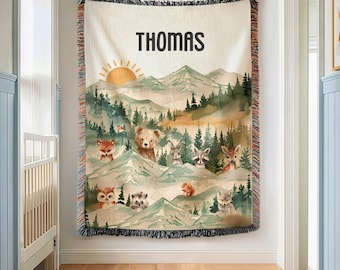 May include: A woven throw blanket featuring a mountain landscape with a sun, pine trees, and various woodland animals including a bear, fox, and deer. The name THOMAS is printed in black at the top. The blanket has a fringed edge on all sides.
