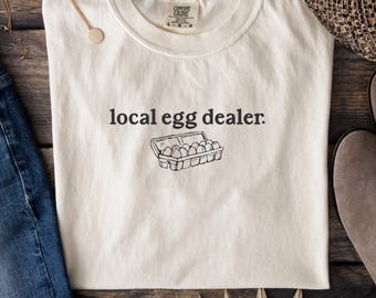 以下が含まれることがあります： 「local egg dealer.」の文字と卵のカートンのイラストがプリントされた、クリーム色のTシャツ。木製の表面に平らに置かれ、金のネックレスとデニムのジーンズが見えます。