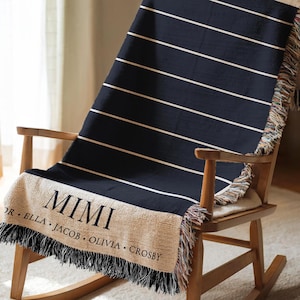 Puede incluir: Una manta a rayas azul marino y blancas con un borde con flecos está sobre una mecedora de madera. La manta presenta el nombre "MIMI" en letras grandes, con nombres adicionales debajo.