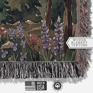 May include: Woven blanket with a forest scene featuring purple lupine flowers, trees, and other wildflowers. The blanket has a fringed edge and the text "WOVEN BLANKET TEXTURE". The blanket is made in the USA and is 100% cotton.