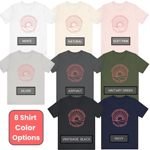 Puede incluir: Se muestran ocho opciones de color de camiseta, que incluyen blanco, natural, rosa suave, plateado, asfalto, verde militar, negro vintage y azul marino. Cada camiseta presenta un dise&ntilde;o gr&aacute;fico de una salida del sol sobre un paisaje con el texto "Every sunrise brings new possibilities" y "The journey is in the pursuit".