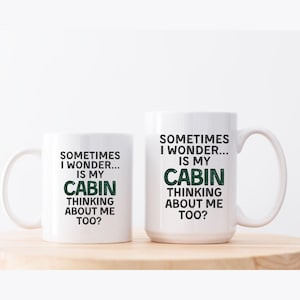 May include: Two white ceramic mugs with the text "SOMETIMES I WONDER... IS MY CABIN THINKING ABOUT ME TOO?" in black and green. The mugs are on a light brown wooden surface.