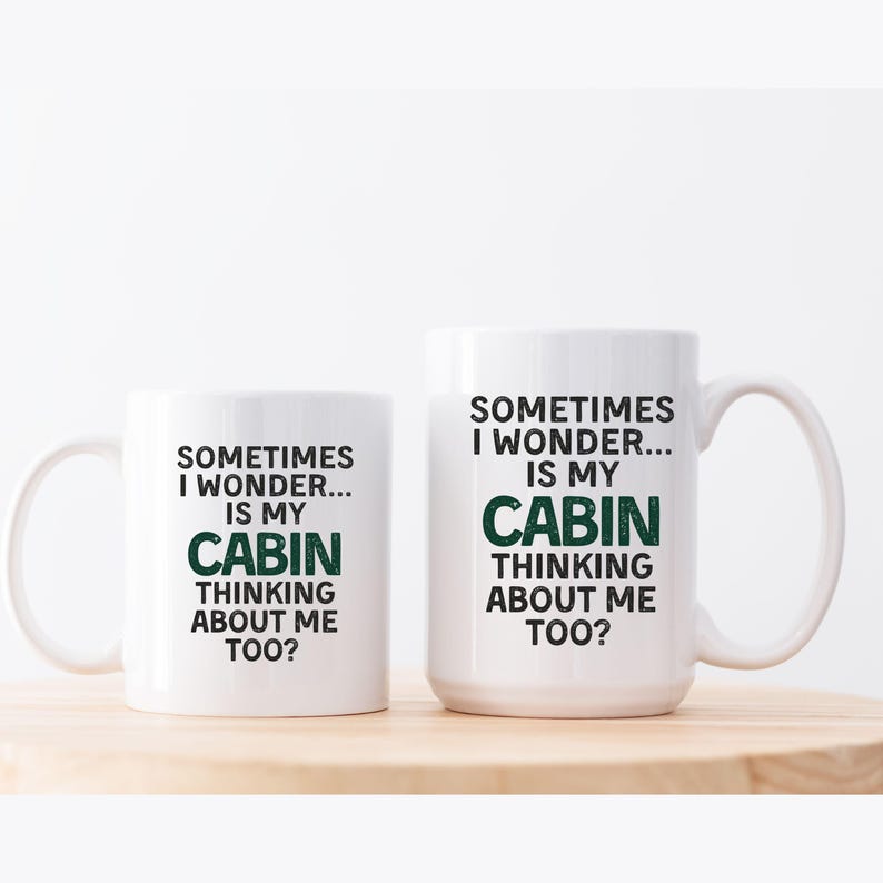 May include: Two white ceramic mugs with the text "SOMETIMES I WONDER... IS MY CABIN THINKING ABOUT ME TOO?" in black and green. The mugs are on a light brown wooden surface.