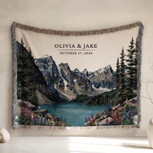May include: A woven throw blanket featuring a watercolour-style mountain lake landscape with pine trees and wildflowers. The text OLIVIA & JAKE and OCTOBER 27, 2026 is printed at the top. The blanket has a fringed edge with a multi-coloured thread design.