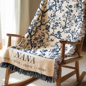 May include: A beige throw blanket with a blue floral pattern is draped over a wooden rocking chair. The blanket has a fringe and the word "NANA" is printed on it, along with the names: Elinor, Ella, Olivia, and Crosby.