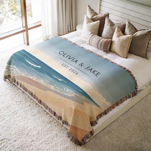 May include: A woven throw blanket featuring a beach scene with blue ocean waves and tan sand. The text OLIVIA & JAKE and EST 2026 is printed in black across the centre. The blanket has a fringed edge and is draped over a bed with several pillows.